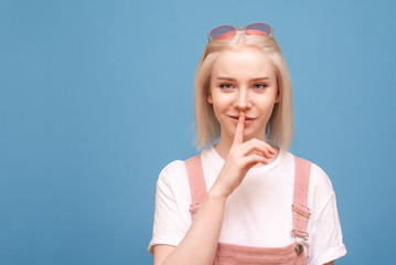 A portrait of a funny teen girl shows a silence sign on a blue background and looks at the camera. Funny girl isolated on a scarlet background, joined the index finger to the lips. Copyspace