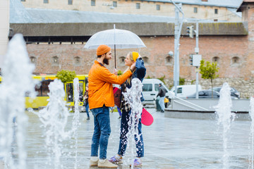 Stylish hipster couple with transparent umbrella in bright casual clothes in city street with a longboard in urban city landscape. Full height of beard man and blonde woman through the fountain water