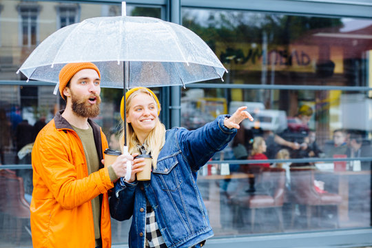 Positive bearded man with transparent umbrella and hiis girlfriend in bright wear pointing by hand looking aside drinks coffee in rainy weather urban city street on background. Lifestyle concept.
