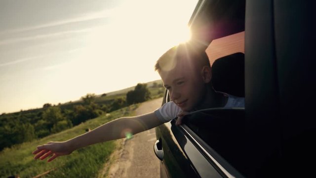 The boy looks out of the car window traveling the world. The boy's face in the sun with an arm stretched out of the car window. The concept of a happy family. Pursuit of travel and entertainment