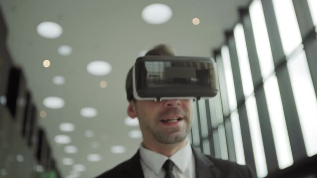 Close-up shot of middle aged businessman in virtual reality headset having video call and talking while walking along office corridor