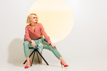 beautiful stylish young woman in colorful clothes sitting in armchair on white with yellow circle