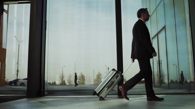 Tracking slow motion shot of elegant businessman in suit walking along airport or hotel hall with transparent glass walls and pulling trolley bag