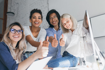 Team of freelance programmers having fun in office and posing with thumbs up. Blissful international students celebrating succesful exams in university and laughing.