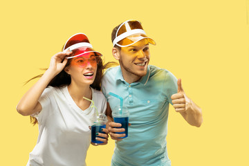 Beautiful young couple's half-length portrait isolated on yellow studio background. Woman and man standing with drinks in colorful caps. Facial expression, summer, weekend concept. Trendy colors.