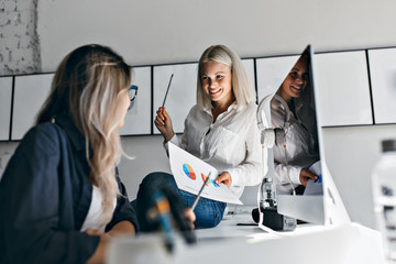 Smiling blonde female manager holding infographic and pencil, while sitting on table. Indoor...