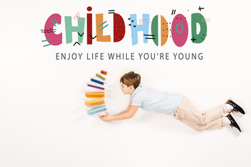 top view of kid holding books and flying near childhood enjoy life while you're young lettering on white