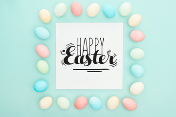 top view of square frame made of painted chicken eggs on blue background with happy Easter black lettering on white greeting card