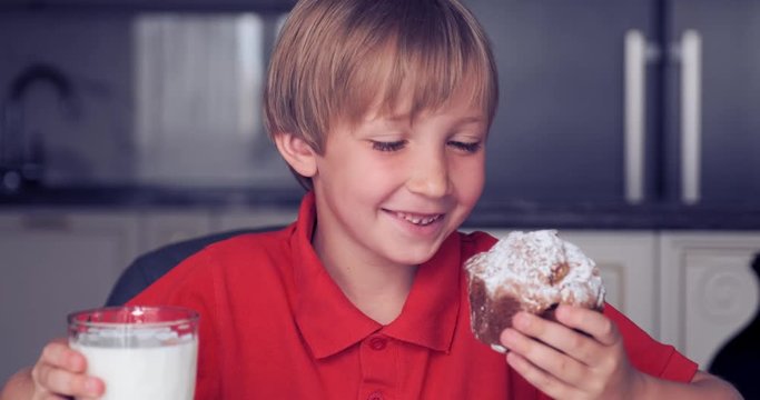 Cute happy kid eats healthy food in the morning.  Little boy drinking milk with cupcake.  7 year old boy having Breakfast with bun and milk.  Blonde boy with a tasty sweet muffin.