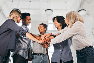Cheerful international students with happy face expression going to work together on science project. Indoor photo of blonde woman in trendy blouse holding hands with coworkers.