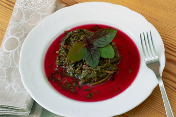 Prepaired braised amaranth - red spinach, in it's own sauce and a fresh leaf on top on white plate on wooden table  with linen napkin and fork nearby, copy space, close up, top view