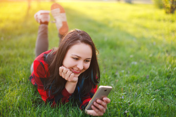 Portrait of a smiling woman lying on green grass and using smartphone outdoors