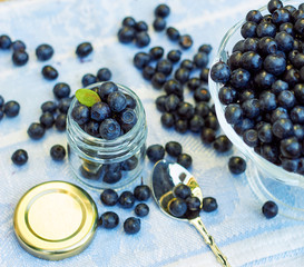 Full jar of fresh whortleberry berries with green leaf on top, with berry spilled around on linen table cloth with spoon and glass vase nearby, copy space, closeup