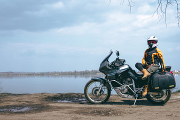 Fototapeta na wymiar Biker girl wearing a motorcycle outfit, protective clothing, equipment, adventure touristic motorbike with side bags. outdoor travel, active traveler, lake and clouds. copy space. dual sport touring
