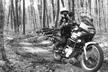 Fototapeta na wymiar Biker girl wearing a motorcycle outfit, protective clothing, equipment, adventure touristic motorbike with side bags. outdoor travel, off road enduro active traveler, sunny forest, black and white