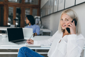 Obraz premium Cute blonde secretary in white blouse talking on phone and writing data in notebook. Indoor portrait of long-haired asian it-specialist with graceful lady at reception desk.