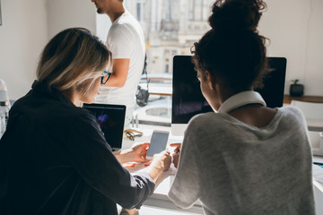 Portrait from back of blonde girl in black shirt sitting beside laptop and showing new phone to...