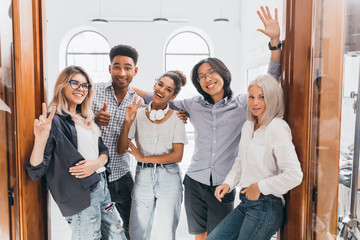 African young man with surprised smile standing beside doors with colleagues after work day. Asian student in glasses posing with university friends near library.