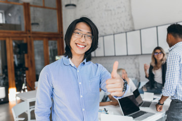 Asian laughing boy posing with thumb up at the beginning of the workday. Chinese office worker in...