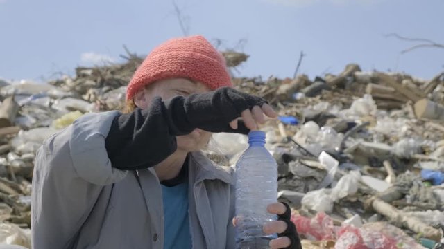 Close-up. homeless woman drinking water from plastic bottle in landfill. bum dwells in the city garbage.