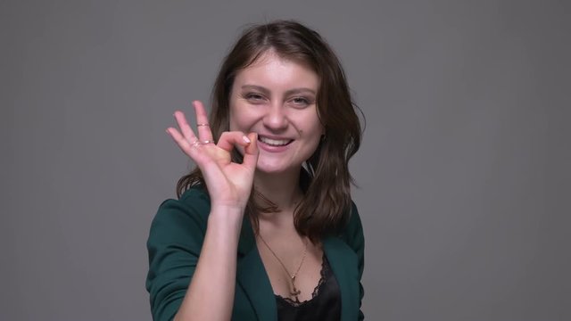 Closeup shoot of adult attractive brunette female showing okay sign smiling and looking at camera with background isolated on gray