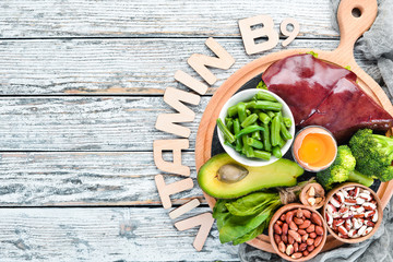 Foods that contain natural vitamin B9: Liver, avocado, broccoli, spinach, parsley, beans, nuts, on a white wooden background. Top view.