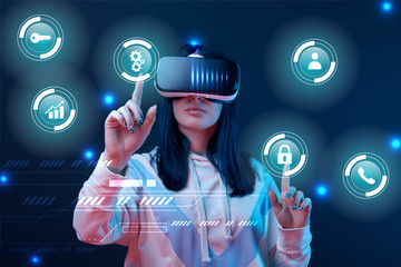 young woman in virtual reality headset pointing with fingers at glowing cyber icons on dark background
