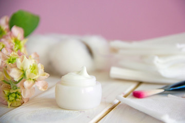A small white open jar with cosmetic white cream stands on a wooden table, next to fresh flowers, cotton napkins, makeup brush. Conception of natural cosmetics. Copy space