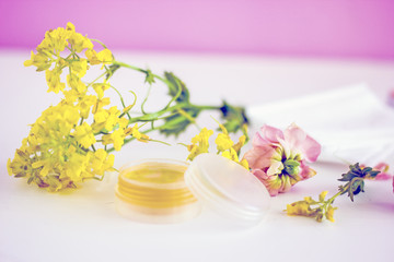  cosmetic oil for the face in a transparent jar with a lid, near wildflowers, twigs, white napkins, pink petals on a white background. Top view 