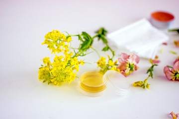  cosmetic oil for the face in a transparent jar with a lid, near wildflowers, twigs, white napkins, pink petals on a white background. Top view 