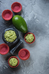 Marble mortar with freshly made guacamole and red vegetable tartlets. Flatlay over grey concrete background, vertical shot, copy space