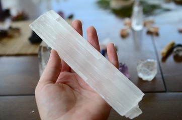 Large Selenite Rod, Holding Crystal over wooden table. Women's hand holding selenite in natural lighting. Healing selenite chunk, large witch wand. Wiccan healing crystal tools, zen meditation stones.