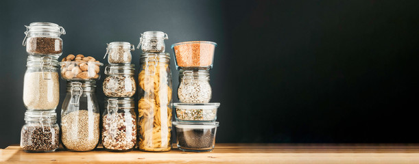 Banner with assortment of uncooked grains, cereals and pasta in glass jars on wooden table. Healthy...