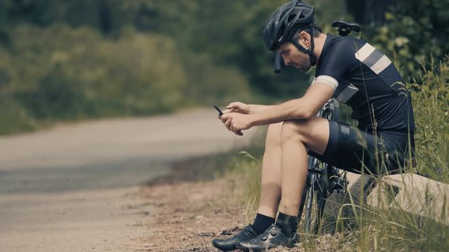 Cyclist Using Smartphone.Athlete Using Smartphone.Handsome Young Sportsman Using Smartphone In Park.Cyclist Lying On Green Grass And Using Mobile Phone.Fit Male Cyclist Watch On Mobile Phone.	