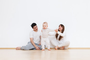 Naklejka premium Positive family portrait with mom,dad and little baby boy standing on white wall background. Parents hold by hand their child and help him do his first step. Generation concept