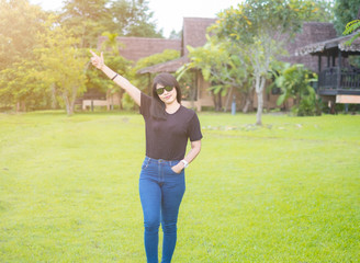 Beautiful Asian Women. wearing sunglasses. Standing poses lift Hands up In a good mood. Wear casual dress black t-shirt with Blue jeans. Green grass. enjoy relax Refreshing. sunlight of the morning.