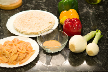 Onions, peppers, chicken meat, tortillas and spices, all the ingredients to do fajitas, a Mexican dish, on a black granite worktop