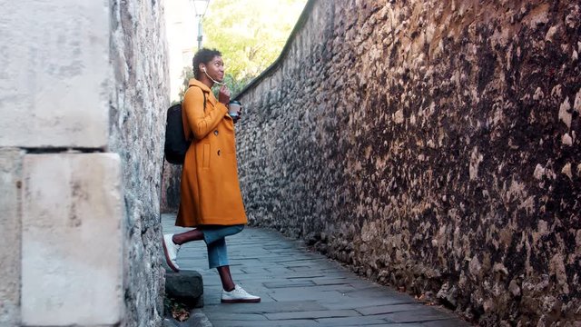 Young woman wearing a yellow pea coat and blue jeans leaning on stone wall in an alleyway talking on her smartphone using earphones, low angle, full length