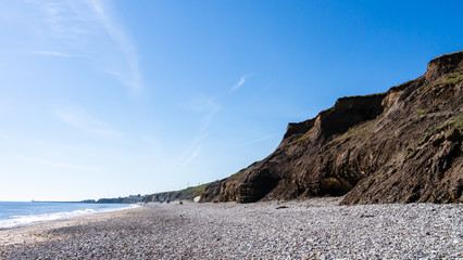 Fototapeta na wymiar Panorama of cliff faces made of rock and earth at Seaham Hall Beach in County Durham showing blue sky, pebbled beach and a calm North Sea.