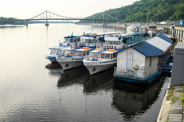 Fototapeta premium Tourist Ships moored on the Dnieper River embankment in Kyiv. Pedestrian bridge on the background. Kyiv, Ukraine. 16-06-2019