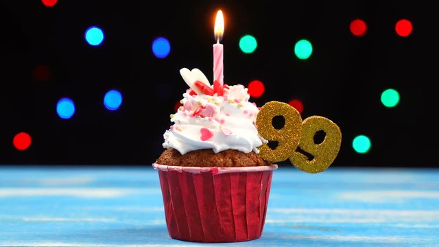 Delicious birthday cupcake with burning candle and number 99 on multicolored blurred lights background