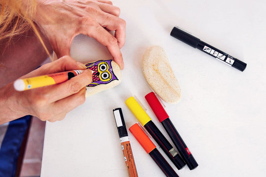 Hands of a woman painting an owl figure on a stone with an acrylic pen. Stone painting handicraft.