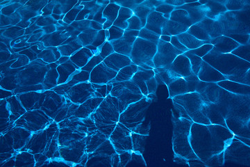 Sun creating abstract dappled light mesh from waves on a blue pool with human shadow