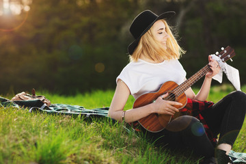 Portrait of a young beautiful woman in a black hat. Girl sitting on the grass and playing guitar