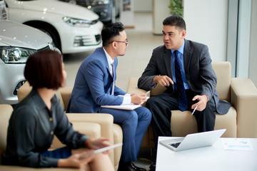 Businessmen sitting on sofa and discussing a purchase of new car with businesswoman sitting near by them and listening to their conversation  in car showroom