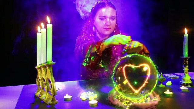 A gypsy woman in a magic salon in puffs of smoke guesses on a crystal ball and a heart sign appears in it.