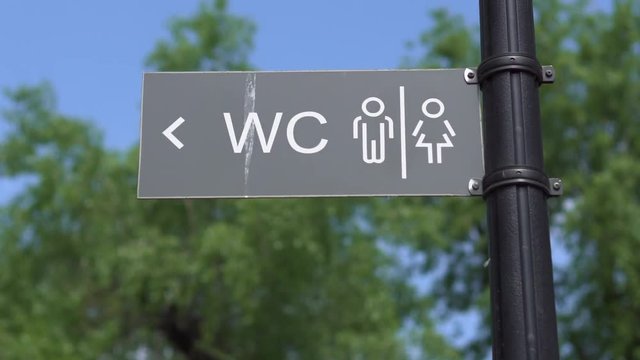 A sign pointer to the toilet outside in a city park against the background of green trees and a blue sky on a sunny day. Close-up