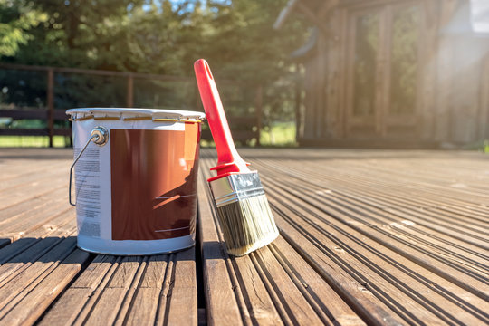 On the painted boards of the terrace there is a can of paint and a large brush. Summer day, the sun illuminates the materials to protect the wood.