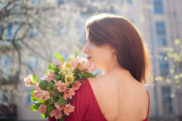 Fototapeta premium Back view of amazing and beautiful young woman holding a big bouquet of colorful flowers outdoors near the buildings on the background
