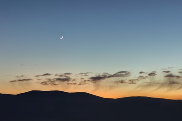 Young moon, mountain silhouettes and beautiful sunset sky with low fluffy clouds